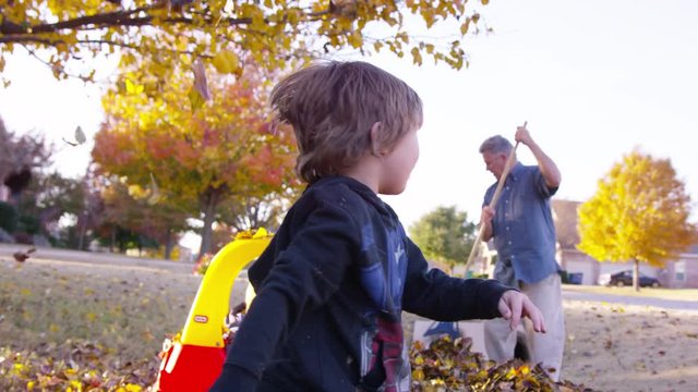 Boy runs to camera and throws leaves at camera while family rakes leaves