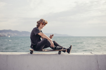 Young man using a cellphone while sitting at longboard on pier witth yacht