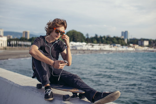 Young man using a cellphone while sitting at longboard on pier witth yacht - Powered by Adobe