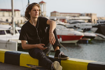 Young man using a cellphone while sitting at longboard on pier witth yacht
