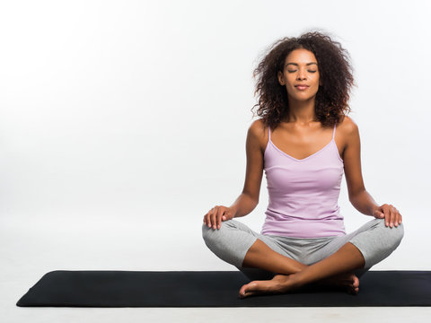 Happy African American Woman In Comfortable Sports Wear Meditating On Mat Over White Wall Background. Black Girl Concentrated On Yoga Practice.