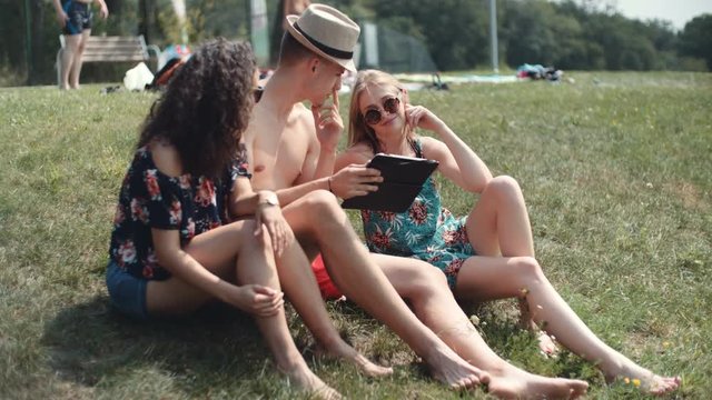 Friends looking on their tablet while sitting on a grass. Happy mixed ethnicity group of friends using computer tablet.