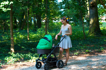 Fototapeta na wymiar Nature walk with stroller, young mother in beautiful dress walking on the forest walkway with her baby in the pram, enjoying fresh air and smiling on her cute newborn, mother and baby stroller walk