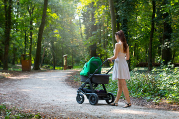 Fototapeta na wymiar Woman in dress walking with the pram, surrounded by beautiful nature, pram walk on the walkway in the forrest park, parenting mother and baby outside during warm summer day