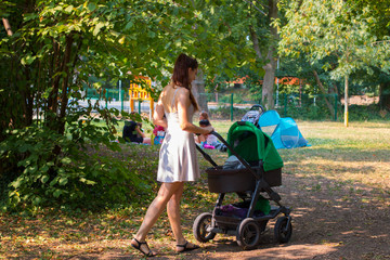 Young woman pram walking in the beautiful park, mother getting out with her baby to walk with her pram, sunny day in baby friendly park, children playground behind, moms sitting on the grass