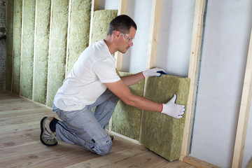 Worker in protective goggles and respirator insulating rock wool insulation in wooden frame for...