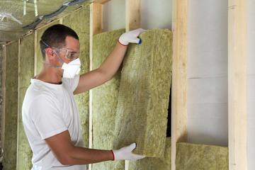 Worker in protective goggles and respirator insulating rock wool insulation in wooden frame for...