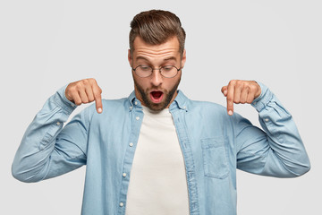 Horizontal shot of surprised unshaven young male points down, opens mouth widely, sees something stunning on floor, wears stylish shirt, isolated over white background. People and emotions concept