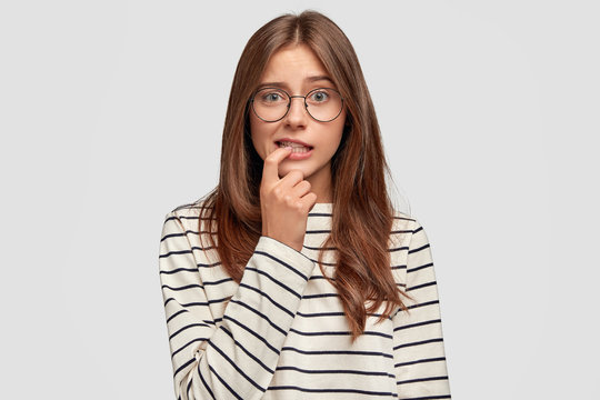 Lovely young woman with thoughtful expression bites finger, dressed in striped sweater, wears round spectacles, poses against white background. Pensive worried female student thinks about something