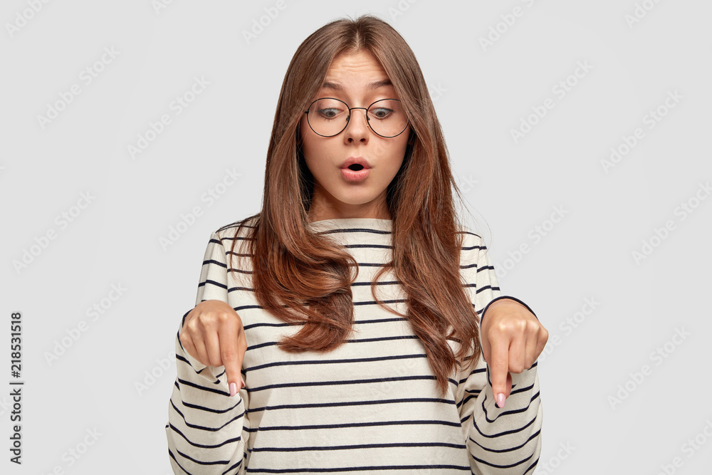 Wall mural Surprised young lovely female teenager opens mouth widely, has startled look, points down with stupefied expression, sees something unbelievable, isolated over white background. Omg, look there!