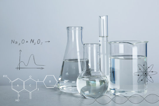 Laboratory glassware with liquids for analysis on table and chemical formulas against light background