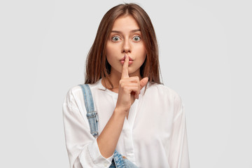Shush, be silent! Serious lovely female asks to keep secret information confidential, dressed in white oversized shirt and denim overalls, poses against white studio wall. Conspiracy concept