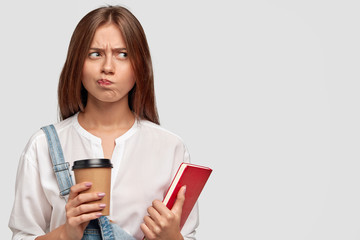 Unhappy lovely female student feels displeased from learning all time, holds cup of takeaway coffee and book, thinks about rest, has sullen expression, isolated over white background with free space