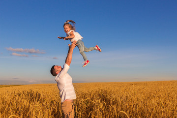Fototapeta premium Happy family - the father throws his daughter in the middle of a wheat field on a sunny summer day. Concept of freedom