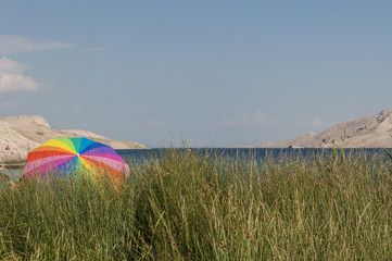 Croazia: ombrellone arcobaleno a Rucica, la spiaggia di ciottoli in una baia brulla con erba, piante verdi e un paesaggio desertico sulla famosa isola di Pago