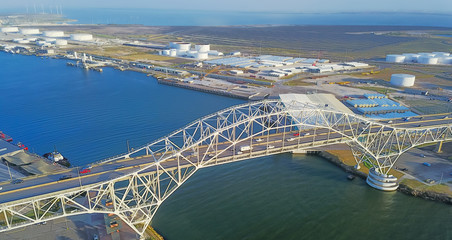 Panorama aerial view of Corpus Christi Harbor Bridge with row of oil tanks and wind turbines farm in distance. A through arch bridge crosses the Corpus Christi Ship Channel