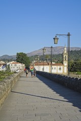 Fototapeta na wymiar Roman bridge in Ponte de Lima, with people on the path. Beautiful historic stone supported by the architecture of its arches. Typical old lamps in Portugal, and the Santo António da Torre church