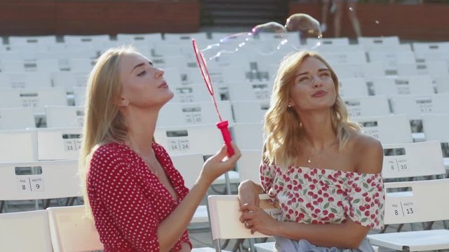 Two attractive beautiful young blond women blow bubbles in the park and are happy on sunset. Girlfriends in park. Slow motion 