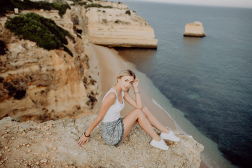 Young blonde girl sitting on the edge of the cliff looking into the ocean. Travel concept.