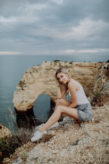 Young blonde Woman sitting on the rocks top by the ocean. Beautiful peace landscape of Portugal beaches.