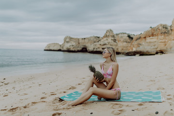 Young slim woman in bikini and sunglasses holding fresh pineapple on the sea beach