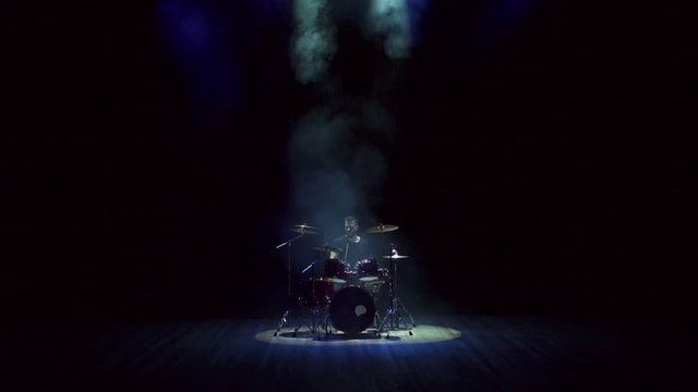 Bearded brutal man plays the drums on a black background on stage with smoke and white light.