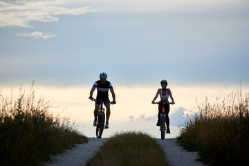 Obraz premium Pair of bicyclist riding bikes on a country road. Fit young people in sportswear cycling downhill against beautiful horizon with perfect sky at evening