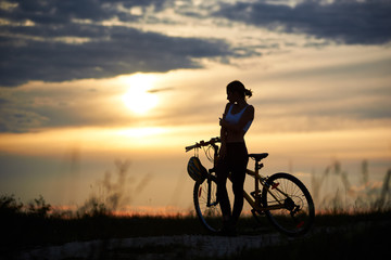 Fototapeta na wymiar Beautiful, slender woman standing near bike and looking at sun and marvelous landscapes. Silhouette of sporty cyclist posing on trail in grass. Concept of motivation and healthy lifestyle.