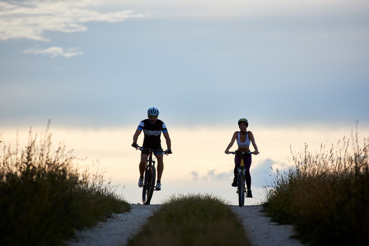 Pair of bicyclist riding bikes on a country road. Fit young people in sportswear cycling downhill against beautiful horizon with perfect sky at evening