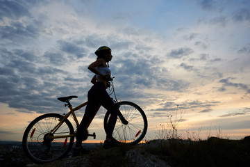 Fototapeta na wymiar Side view athletic girl in helmet with bicycle on rock under beautiful evening sky with clouds on background of city in the distance. Copy space