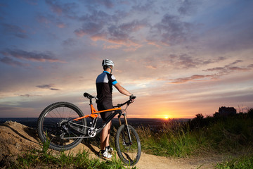 Fototapeta na wymiar Back view of cyclist male in a helmet and sports clothes rests on the background of a sunset from a height watching the mountain in the distance, the enchanting sky and the bright sun