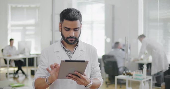 Young attractive surgeon standing and using iPad. Studying pictures and diagnosis of the patient. In background medical clinic and working doctors
