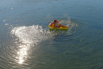 Fototapeta premium A beautiful young woman relaxes on an inflatable sun bed in the middle of a lake. She wears a white swim suit and enjoys the summer sun setting as she leisures on her sun bed. 