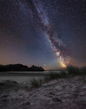 Vibrant Milky Way composite image over landscape of yellow sandy beach Three Cliffs bay