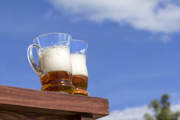 Two muggs of beer are on the table against the sky and clouds
