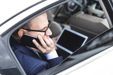 Young businessman talking on mobile phone and using tablet pc while sitting on back seat of a car. Caucasian male business executive by a taxi and looking at digital tablet.