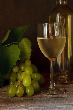Green grapes with leaves, glass, bottle of white wine on vintage wooden background. Close-up, selective focus. Still life in vintage stile. Dark photo.