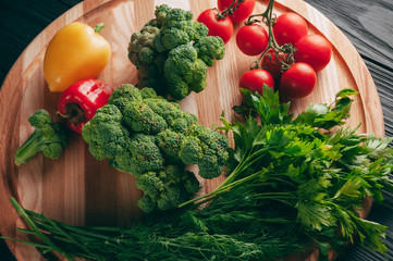 Fresh vegetables: cherry tomatoes, broccoli, pepper, dill, parsley on a round wooden board, on a dark wooden table. Recipe. Ingredients. Dietary food. Place under the text. View from above