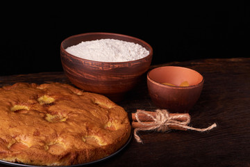 On a dark wooden table, fashionable bakeries, a bowl with flour, apricot cake and tools stand on table