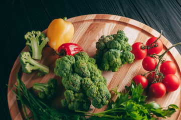 Fresh vegetables: cherry tomatoes, broccoli, pepper, dill, parsley on a round wooden board, on a dark wooden table. Recipe. Ingredients. Dietary food. Place under the text. View from above