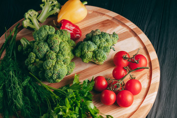 Fresh vegetables: cherry tomatoes, broccoli, pepper, dill, parsley on a round wooden board, on a dark wooden table. Recipe. Ingredients. Dietary food. Place under the text. View from above
