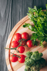 On a dark wooden table, on a round wooden board fresh green broccoli, parsley, dill and cherry tomatoes for your health. Recipe. Ingredients. Dietary food. Place under the text. View from above