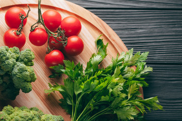 On a dark wooden table, on a round wooden board fresh green broccoli, parsley, dill and cherry tomatoes for your health. Recipe. Ingredients. Dietary food. Place under the text. View from above