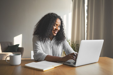 Gorgeous positive young dark skinned female blogger keyboarding on generic laptop, smiling, being...