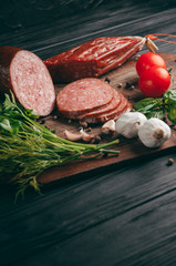 Smoked salami on a black table and a brown board. Sausages with rosemary, garlic and pepper and tomato cherry, dill, parsley.