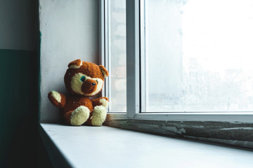 Abandoned children's toy teddy bear unhappy child sits on the sills alone in the entrance and looks out the window