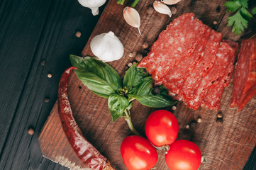 Smoked salami on a black table and a brown board. Sausages with rosemary, garlic and pepper and tomato cherry, dill, parsley