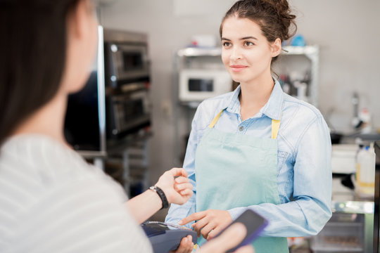 Waist up of smiling young woman wearing apron looking at customer paying via NFC in shop, copy space