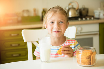 Cute girl child eats cookies and drinks milk sitting in the kitchen