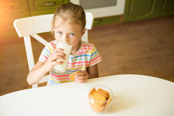 Cute girl child eats cookies and drinks milk sitting in the kitchen
